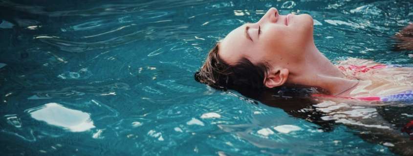 A smiling woman floating on her back in a swimming pool. Self-care is one way to manage stress effectively.