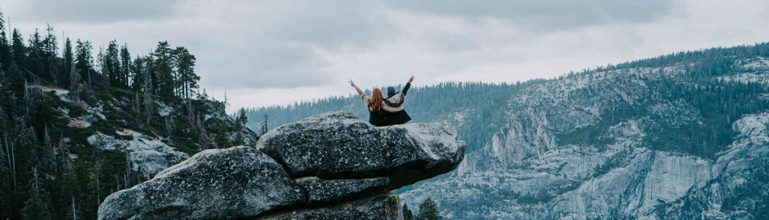 Two people sitting close to one another on a rock precipice with their arms stretched in the air with joy.