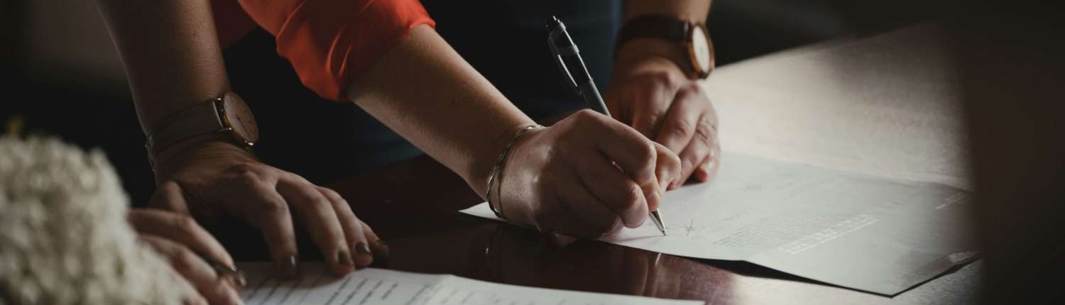 A woman in an orange shirt filling out paperwork on a wood table. Another person is looking over it.