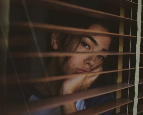 Socially Anxious woman looking through blinds wishing she was outside with other friends.