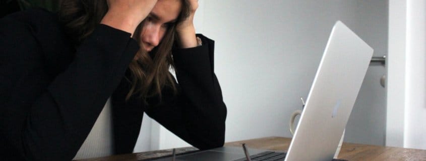 A woman in a black shirt holds her head in exasperation while looking at work on her laptop.