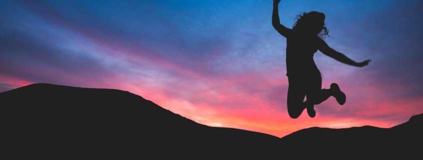 A silhouette of a woman jumping in front of mountains and a colorful sky.