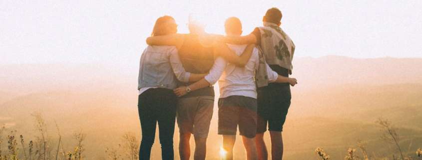 A group of four friends standing arm-in-arm against a beautiful sunset with a backdrop of mountains.
